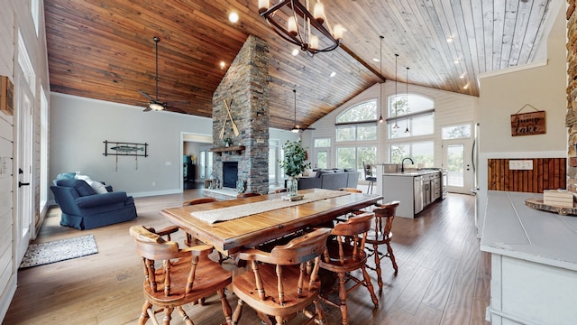 dining space with ceiling fan with notable chandelier, high vaulted ceiling, light wood-type flooring, wooden ceiling, and a stone fireplace