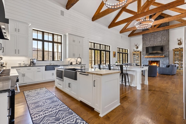 kitchen with an inviting chandelier, beamed ceiling, a kitchen island with sink, hardwood / wood-style floors, and a stone fireplace