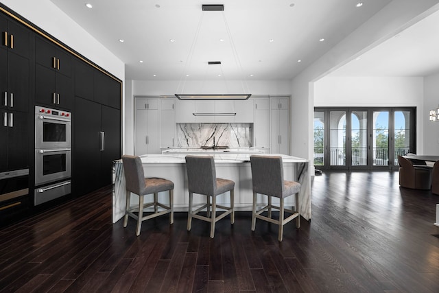 kitchen with backsplash, hanging light fixtures, double oven, wood-type flooring, and a large island with sink