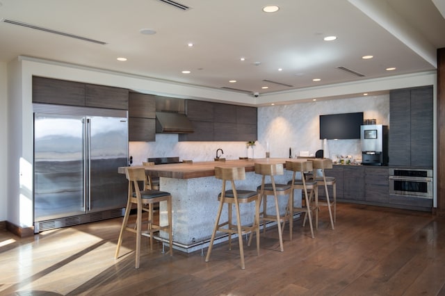 kitchen featuring wall chimney exhaust hood, tasteful backsplash, a breakfast bar area, dark wood-type flooring, and stainless steel appliances