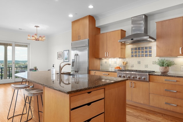 kitchen featuring wall chimney exhaust hood, light hardwood / wood-style floors, pendant lighting, a center island with sink, and appliances with stainless steel finishes