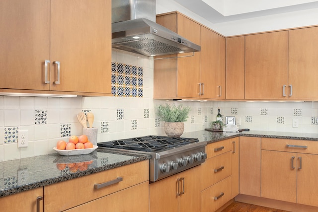 kitchen featuring stainless steel gas stovetop, wall chimney range hood, backsplash, and dark stone counters