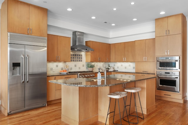 kitchen featuring a center island with sink, wall chimney exhaust hood, stainless steel appliances, and light wood-type flooring