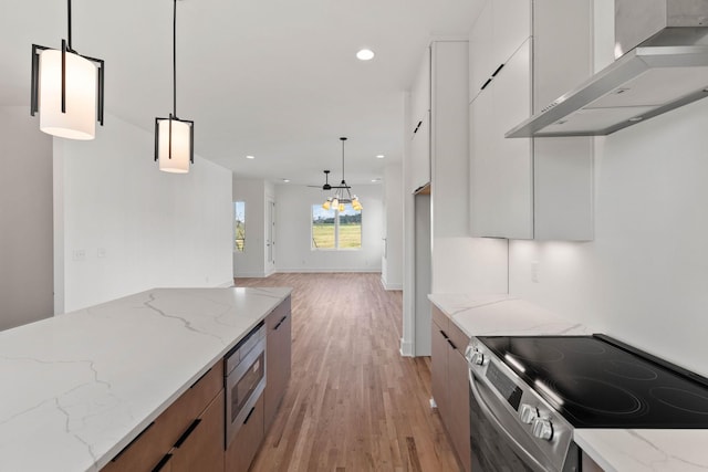 kitchen with light stone counters, stainless steel appliances, wall chimney range hood, white cabinetry, and hanging light fixtures