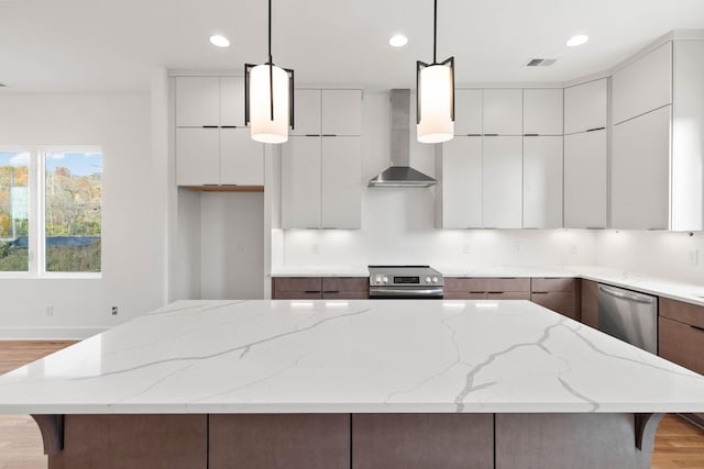 kitchen featuring a center island, wall chimney range hood, appliances with stainless steel finishes, decorative light fixtures, and light hardwood / wood-style floors