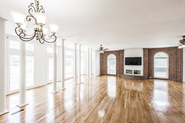 unfurnished living room featuring ceiling fan with notable chandelier, light hardwood / wood-style floors, brick wall, and ornate columns