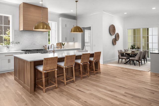 kitchen featuring decorative light fixtures, backsplash, a center island with sink, white cabinetry, and light wood-type flooring