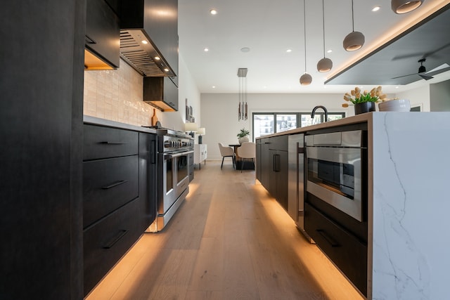 kitchen with light hardwood / wood-style flooring, tasteful backsplash, ceiling fan, wall chimney exhaust hood, and pendant lighting