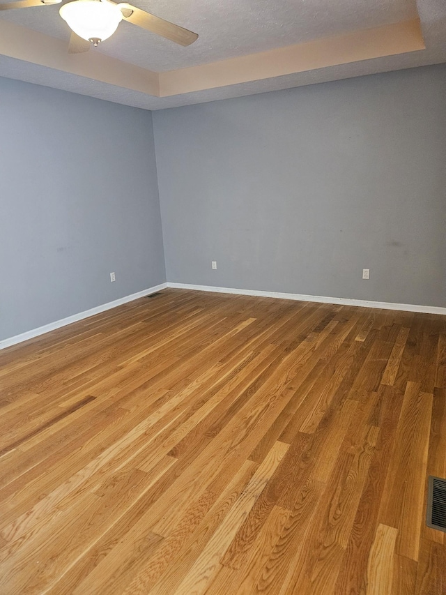 empty room with ceiling fan, wood finished floors, visible vents, baseboards, and a tray ceiling