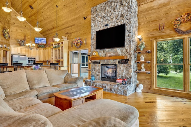 living room featuring a chandelier, wood walls, light hardwood / wood-style floors, high vaulted ceiling, and a stone fireplace