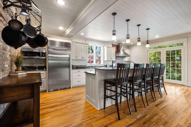 kitchen with wall chimney range hood, white cabinetry, hanging light fixtures, stainless steel appliances, and a kitchen island