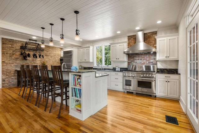 kitchen featuring pendant lighting, wall chimney range hood, high end appliances, an island with sink, and white cabinets