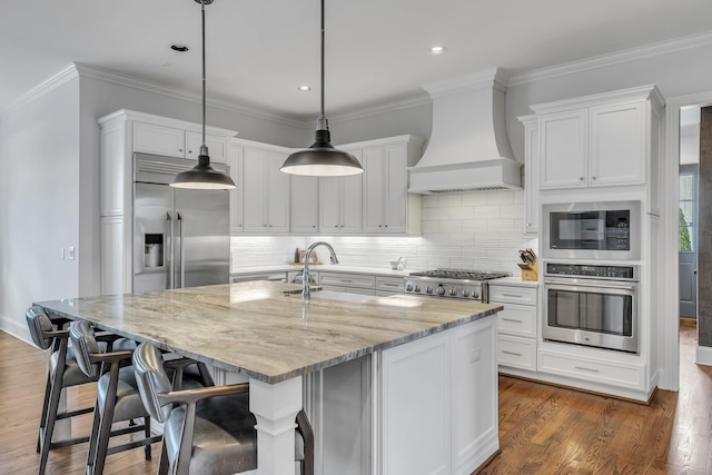 kitchen with light stone counters, built in appliances, pendant lighting, a center island with sink, and custom range hood