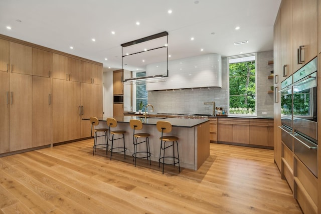 kitchen featuring a breakfast bar, an island with sink, oven, hanging light fixtures, and light wood-type flooring