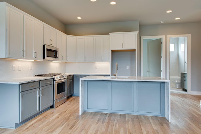 kitchen with sink, a kitchen island with sink, stainless steel appliances, light hardwood / wood-style floors, and white cabinets