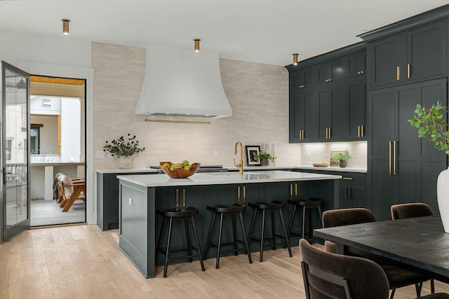 kitchen featuring backsplash, custom exhaust hood, a kitchen island with sink, and light hardwood / wood-style flooring