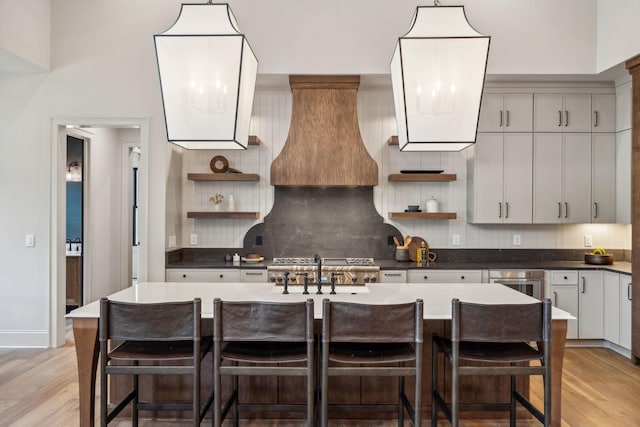 kitchen featuring backsplash, premium range hood, a kitchen island with sink, sink, and light wood-type flooring