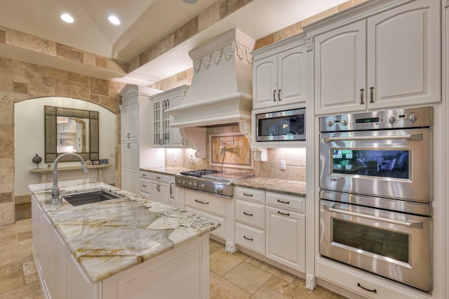 kitchen with stone tile floors, custom range hood, appliances with stainless steel finishes, light stone counters, and a sink