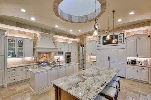 kitchen featuring stone tile floors, premium range hood, a spacious island, a sink, and glass insert cabinets