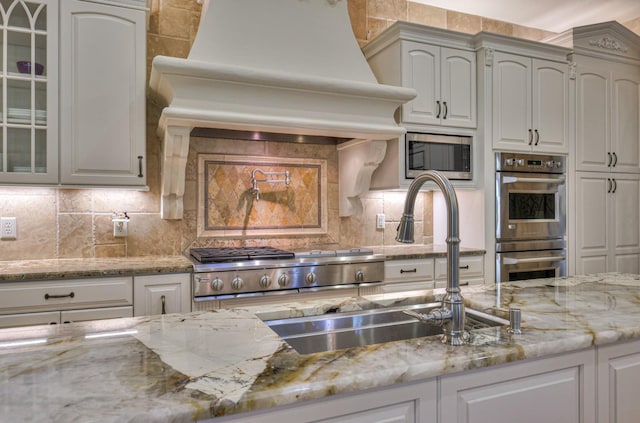 kitchen featuring tasteful backsplash, custom range hood, glass insert cabinets, appliances with stainless steel finishes, and a sink