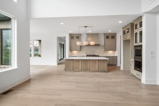 kitchen featuring pendant lighting, double oven, tasteful backsplash, a center island with sink, and light wood-type flooring