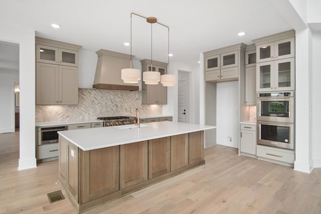 kitchen with double oven, premium range hood, light countertops, an island with sink, and glass insert cabinets