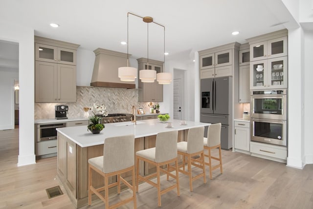 kitchen featuring light countertops, appliances with stainless steel finishes, custom exhaust hood, an island with sink, and glass insert cabinets