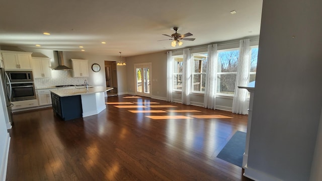 kitchen featuring a center island with sink, wall chimney exhaust hood, appliances with stainless steel finishes, decorative light fixtures, and white cabinetry