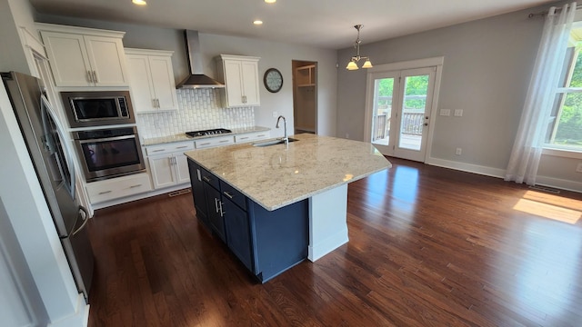 kitchen with white cabinets, appliances with stainless steel finishes, an island with sink, and wall chimney range hood