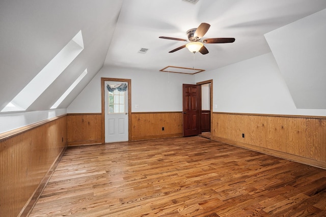 additional living space featuring vaulted ceiling with skylight, ceiling fan, wooden walls, and light hardwood / wood-style flooring