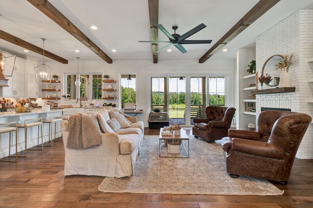 living room with dark hardwood / wood-style flooring, a brick fireplace, ceiling fan with notable chandelier, sink, and beamed ceiling