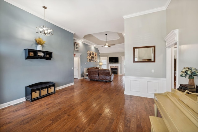 living room with ceiling fan with notable chandelier, a raised ceiling, crown molding, dark wood-type flooring, and a tile fireplace