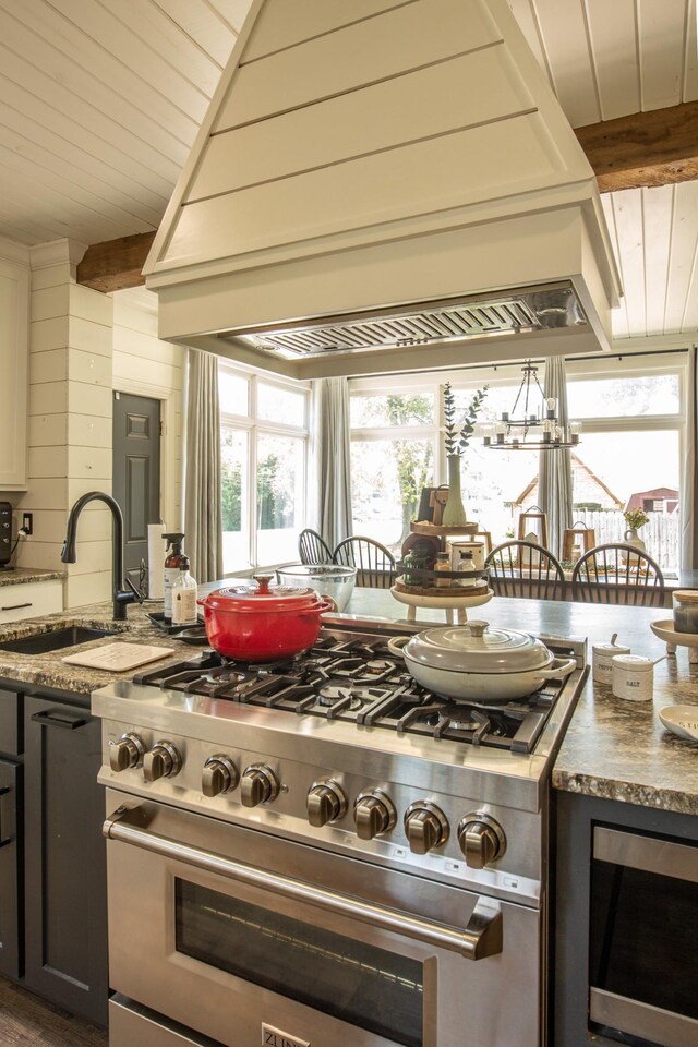 kitchen with stainless steel stove, dark stone counters, custom range hood, sink, and lofted ceiling with beams