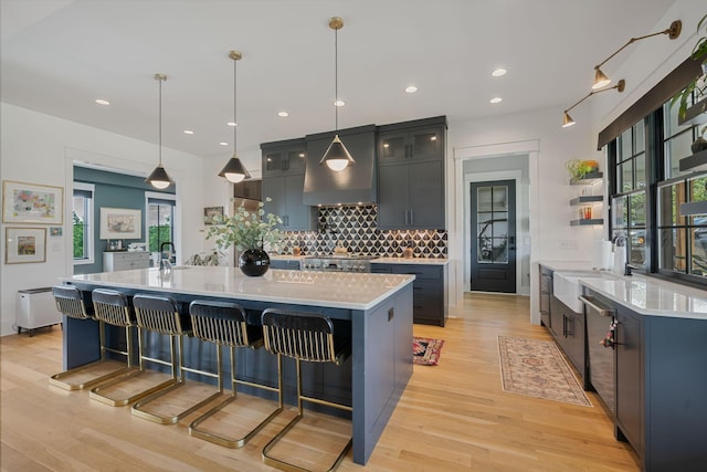 kitchen with hanging light fixtures, a kitchen island with sink, light wood-type flooring, and wall chimney exhaust hood