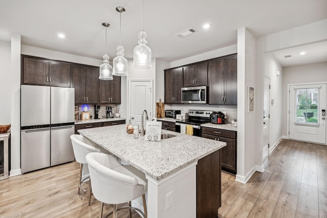 kitchen featuring a kitchen island with sink, light wood-type flooring, hanging light fixtures, and appliances with stainless steel finishes