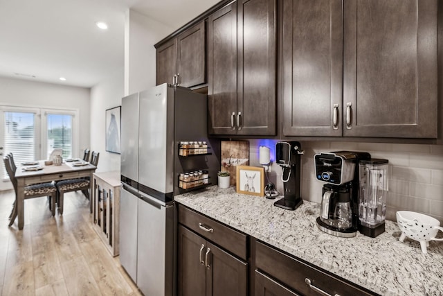 kitchen featuring decorative backsplash, light wood-type flooring, dark brown cabinets, light stone counters, and stainless steel refrigerator