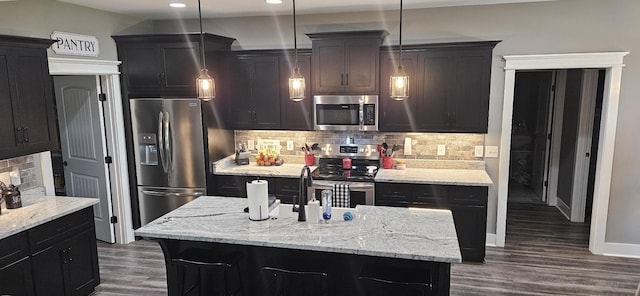 kitchen with light stone countertops, backsplash, stainless steel appliances, hanging light fixtures, and an island with sink