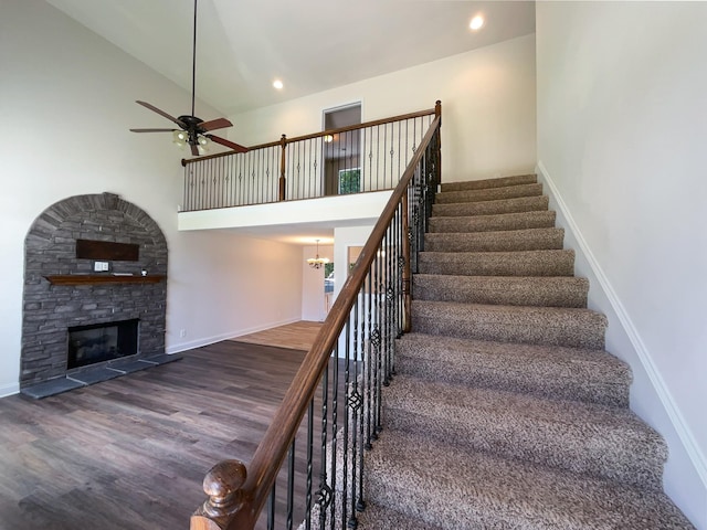 staircase with hardwood / wood-style floors, ceiling fan with notable chandelier, a stone fireplace, and a high ceiling