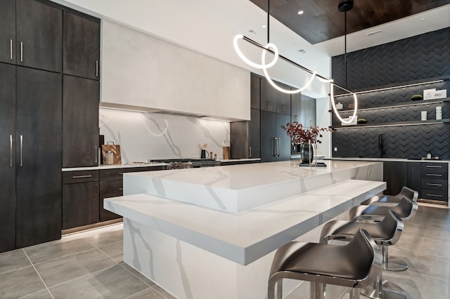 kitchen with a breakfast bar area, a center island with sink, light stone countertops, dark brown cabinetry, and decorative light fixtures