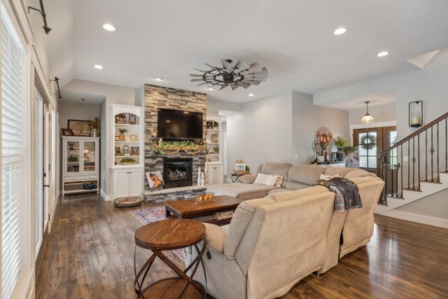 living room featuring a stone fireplace, dark wood-type flooring, built in shelves, and ceiling fan