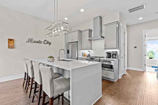 kitchen featuring a breakfast bar, gray cabinetry, a kitchen island with sink, stainless steel appliances, and wall chimney range hood