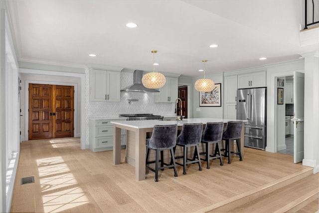 kitchen featuring pendant lighting, high end refrigerator, a center island with sink, a kitchen breakfast bar, and wall chimney range hood