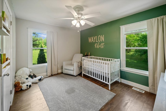 bedroom featuring ceiling fan, multiple windows, dark hardwood / wood-style floors, and a crib