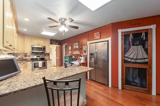 kitchen with light stone countertops, stainless steel appliances, kitchen peninsula, cream cabinets, and ceiling fan