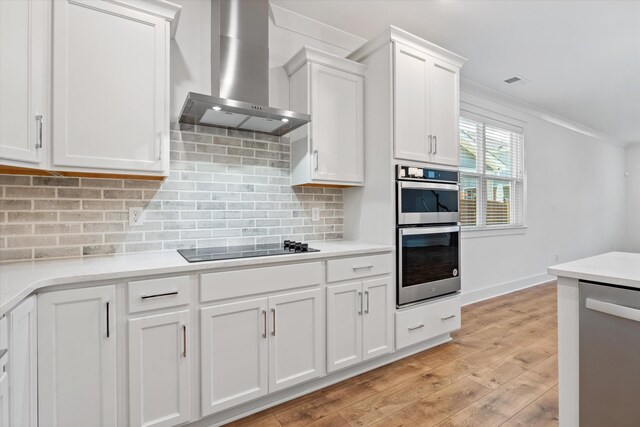 kitchen with wall chimney range hood, light hardwood / wood-style flooring, stainless steel appliances, ornamental molding, and white cabinets