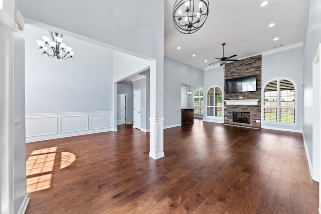 unfurnished living room featuring crown molding, ceiling fan with notable chandelier, dark hardwood / wood-style flooring, ornate columns, and a stone fireplace