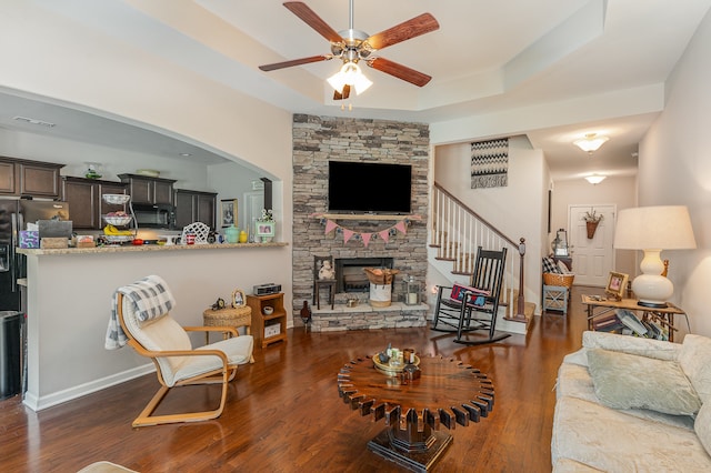 living room with dark hardwood / wood-style floors, a stone fireplace, a raised ceiling, and ceiling fan