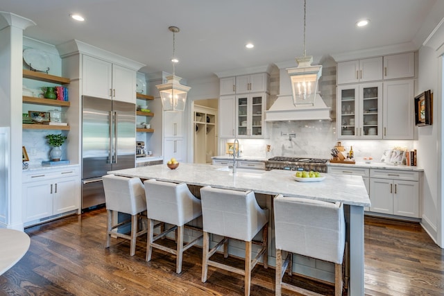 kitchen featuring custom range hood, white cabinets, a kitchen bar, an island with sink, and built in fridge