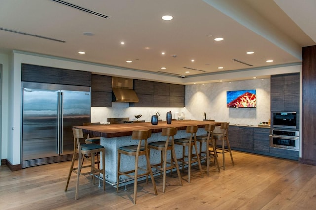 kitchen featuring wood counters, stainless steel built in refrigerator, light wood-type flooring, a kitchen breakfast bar, and wall chimney range hood