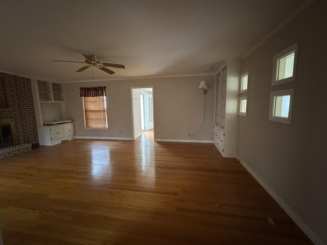 unfurnished living room featuring built in shelves, ceiling fan, a brick fireplace, crown molding, and wood-type flooring